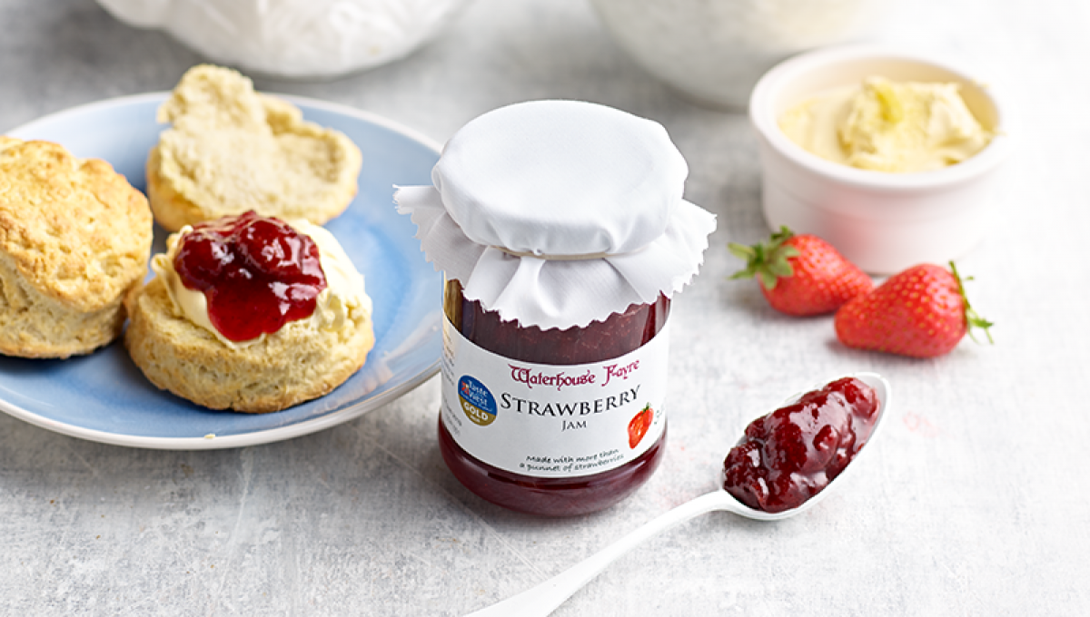 Jar of Waterhouse Fayre strawberry jam next to scones with cream and jam and some strawberries