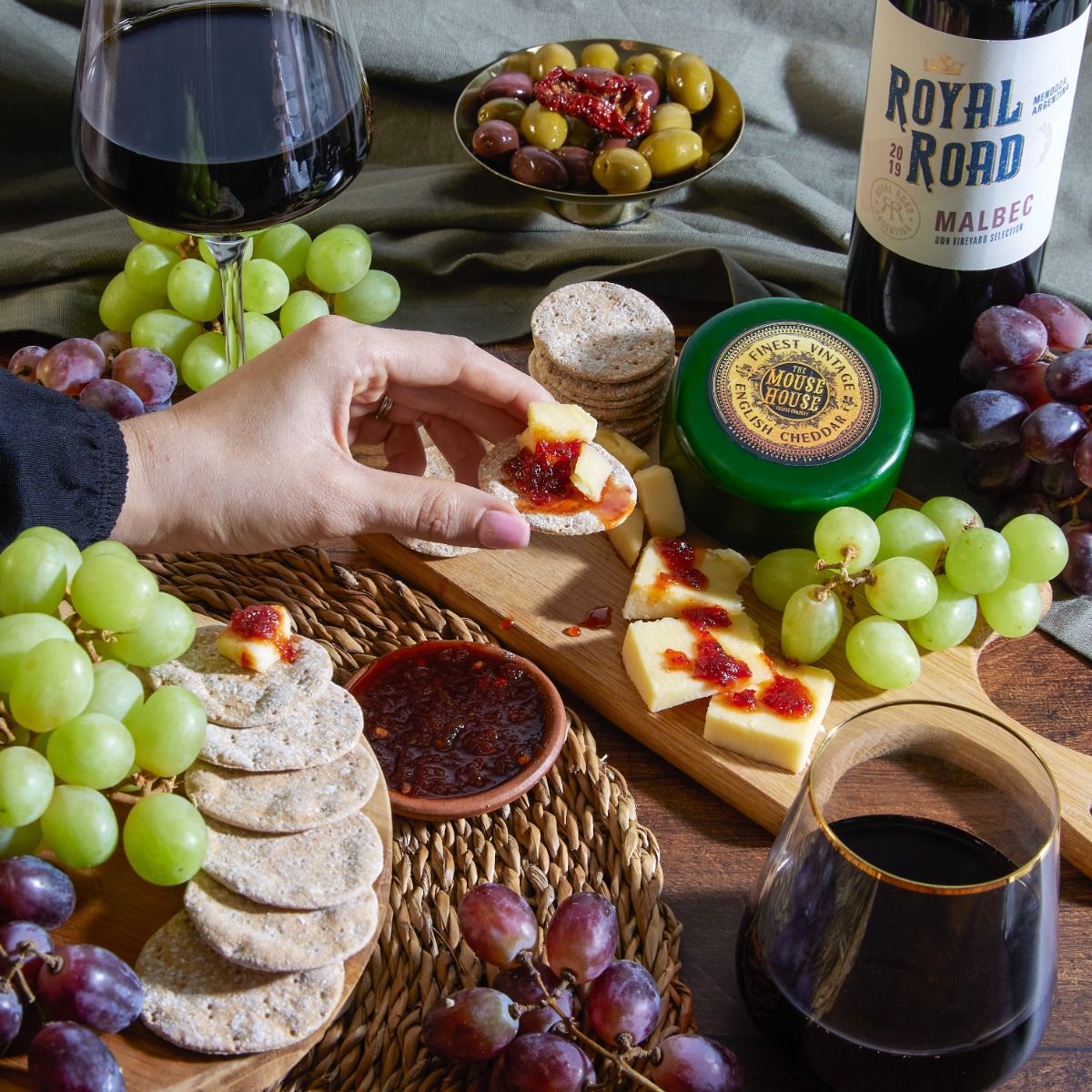 Hand reaching in to eat cheese near an array of cheeses, accompaniments and red wine